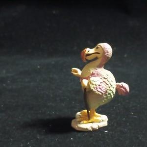 May include: A small, hand-painted figurine of a Dodo bird. The bird has a cream-colored body with pink and green accents on its wings and tail. It stands on a small, beige base with yellow feet and a red beak.