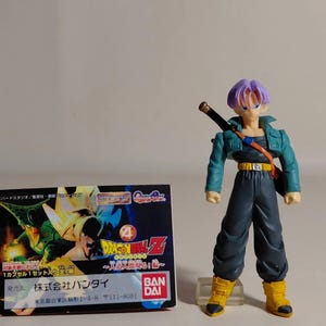 May include: A Dragon Ball Z action figure of a character with purple hair, a teal jacket, and dark pants, holding a sword. The figure stands next to a box with Japanese text and Dragon Ball Z graphics.