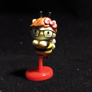 以下が含まれることがあります： ミツバチをテーマにしたデザインのキャラクターのフィギュア。キャラクターは白い顔、黒いメガネ、赤いリボンをしています。体は黄色と黒のストライプで、小さな白い羽が付いています。赤い台座の上に立っています。