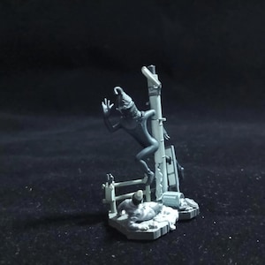 May include: A grey miniature sculpture of a creature climbing a structure. The figure has a long, pointed head and is posed mid-climb. The structure includes a ladder and other architectural details, all in a monochromatic grey.