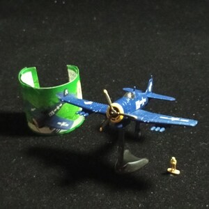 May include: A blue toy airplane with a yellow propeller and white markings, mounted on a black stand. A small gold figurine and a green and white paper cylinder are also present. The airplane has a wingspan of approximately 3 inches.