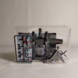 Puede incluir: Un kit de modelo de avión en un embalaje de plástico transparente. El avión sin montar es principalmente gris oscuro con "USAF" impreso en el lateral. Incluye un bebedero de piezas pequeñas y una hélice separada.