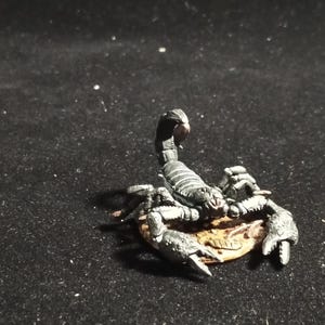 Puede incluir: Una detallada figura de escorpión en miniatura en tonos grises y negros, con cola curvada y pinzas detalladas. El escorpión descansa sobre una pequeña base de color marrón claro, sobre un fondo oscuro.