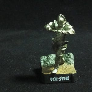 Puede incluir: Figura dorada de un guerrero cyborg en pose dinámica, de pie sobre una pequeña base con superficie texturizada. La base tiene texto japonés. El cyborg lleva una armadura detallada.