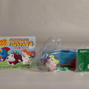 Op de afbeelding: Een verzameling Hamtaro capsule speelgoed, met een trein, een bruine hamster en een witte hamster met oranje oren. De doos heeft Japanse tekst en de woorden "GO GO!" in geel. Ook inbegrepen zijn wielstickers.