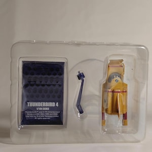 Può includere: Un modello di Thunderbird 4 giallo e rosso in una confezione di plastica trasparente. Il modello è in scala 1/144. Un supporto blu e una base rettangolare blu con testo sono anche nella confezione. Il testo sulla base recita "THUNDERBIRD 4 1/144 SCALE".