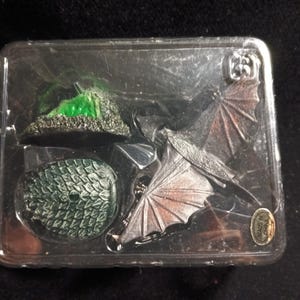 May include: A miniature dragon figurine set in a clear plastic package. The set includes a dragon, a green and black landscape, and a green dragon egg. The dragon has silver wings and a dark body.