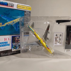 以下が含まれることがあります： 透明なプラスチックケースに入った模型飛行機。飛行機は黄色とグレーで、カラフルなデザインです。パッケージには、「1/700 SCALE」や「MOVIE ARCHIVE」など、日本語と英語のテキストが含まれています。