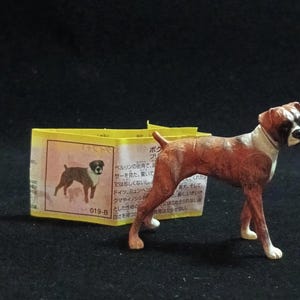 May include: A small Boxer dog figurine, brown and white, on a dark surface. Behind it is a yellow tag with Japanese text and a Boxer illustration. The figurine is a detailed model.