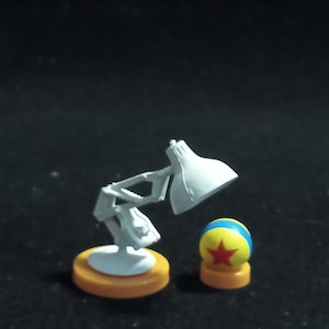 Può includere: Una lampada da scrivania grigia in miniatura con un paralume bianco e una base gialla. Accanto c'è una pallina con una stella rossa, strisce blu e gialle e una base gialla. Gli oggetti sono su sfondo scuro.