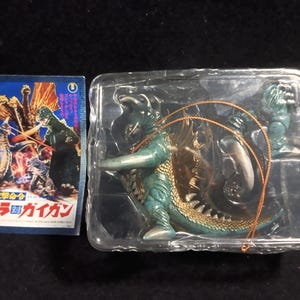 Könnte beinhalten: Eine Godzilla Gigan Actionfigur in einer transparenten Kunststoffverpackung, mit einem japanischen Filmplakat. Die Figur ist teal und goldfarben, mit einem detaillierten Design. Das Poster zeigt eine farbenfrohe Illustration von Godzilla und anderen Monstern.