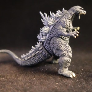 May include: A gray Godzilla figurine with white dorsal fins and a long tail. The toy is posed in a walking stance, with its mouth open. The figure has textured skin and is set against a dark background.