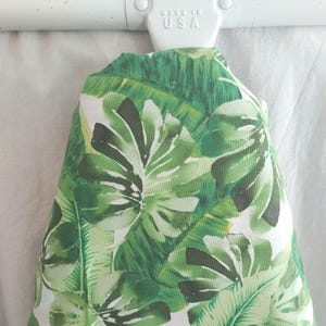May include: A tropical-themed fabric item with a vibrant green and white leaf pattern. The fabric is draped over a white rod, with a "MADE IN USA" marking visible. The design features large, detailed leaves.