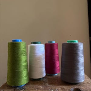 May include: Four spools of thread in green, white, burgundy, and grey. The spools are sitting on a wooden surface.