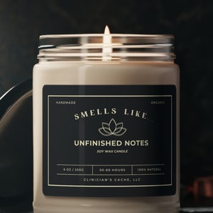 May include: A black and gold soy wax candle jar with the text "Smells Like Unfinished Notes" and "Clinician's Cache, LLC". The candle is lit and the jar is on a dark surface.