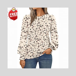 Può includere: Camicetta a maniche lunghe color crema con un motivo a pois neri, rossi e beige. La camicetta presenta un davanti plissettato, polsini arricciati e uno scollo tondo. La modella indossa jeans blu.