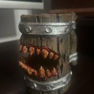 May include: A wooden mug with a monster mouth design. The mug is brown with silver bands and features sharp, yellow teeth with red accents. The open mouth design gives the mug a unique, fantasy-themed appearance.