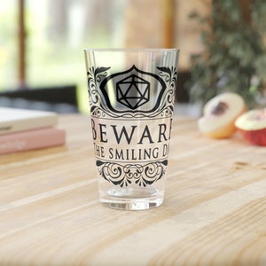 Puede incluir: Un vaso de pinta de vidrio transparente con un diseño negro que presenta un dado estilizado y el texto "Beware the Smiling Dice".