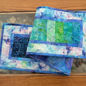 May include: A rectangular quilted table runner featuring a patchwork design with shades of blue, teal, and green. The fabric displays a mix of mottled, floral, and dotted patterns with a dark blue center panel and a quilted texture throughout.