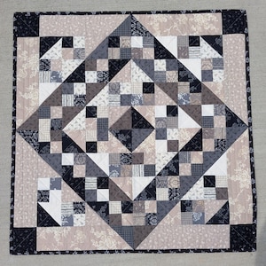 May include: A square quilt featuring a geometric patchwork design in shades of black, white, gray, and beige. The pattern consists of small squares and triangles arranged in a central diamond motif, surrounded by a floral-patterned border.
