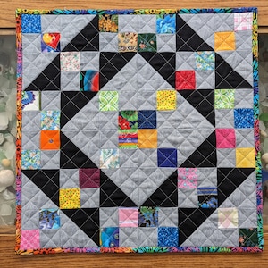 May include: A small quilt with a geometric pattern of black, white, and colorful squares. The quilt is made with a variety of fabrics and has a gray background.
