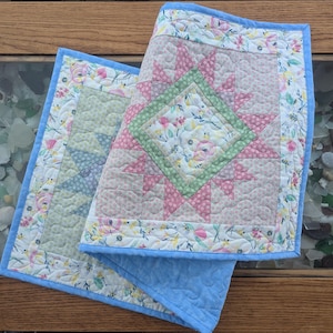 May include: Two quilted table runners with floral patterns. One runner is light blue with a white floral pattern and the other is white with a pink, green, and white floral pattern.