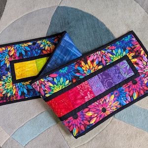 May include: Two colorful, rectangular quilted placemats with a floral design. The placemats feature a black border and a vibrant mix of colors, including pink, blue, yellow, and green. One placemat has a yellow center, while the other has a patchwork center.