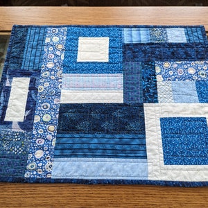 May include: A blue and white patchwork quilt placemat with a geometric design. The placemat is made of various shades of blue fabric with white accents.