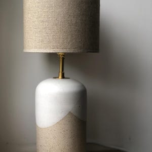 Peut inclure: Une lampe de table en céramique avec un glaçage blanc et beige. La lampe a une base cylindrique et un abat-jour en lin.