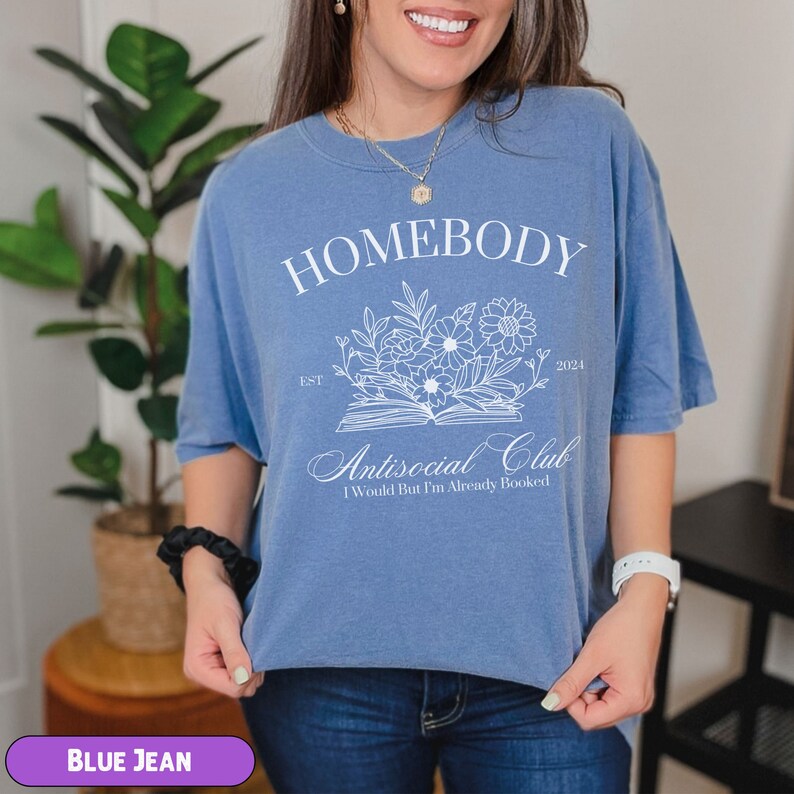 May include: A blue jean t-shirt with white text that reads "Homebody Est. 2021 Antisocial Club I Would But I'm Already Booked". The text is surrounded by a floral design.