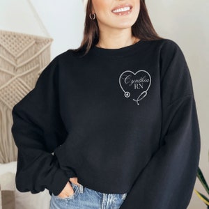 K&ouml;nnte beinhalten: Schwarzer Pullover mit einem wei&szlig;en Herzdesign und dem Text "Cynthia RN" im Herzen. Ein Stethoskop ist um das Herz gezeichnet.