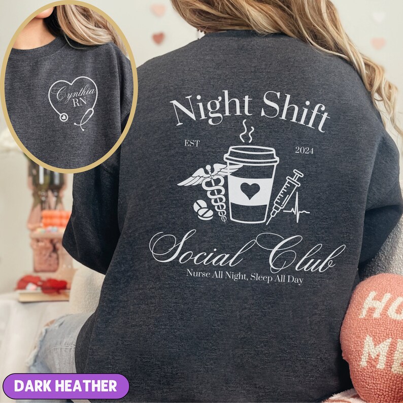 K&ouml;nnte beinhalten: Dunkelgrauer Sweatshirt mit einem wei&szlig;en Grafikdesign. Das Design zeigt eine Kaffeetasse mit einem Herzen, einem Stethoskop, einer Spritze und Pillen. Der Text "Night Shift Est 2024 Social Club Nurse All Night, Sleep All Day" ist auf dem Sweatshirt aufgedruckt.