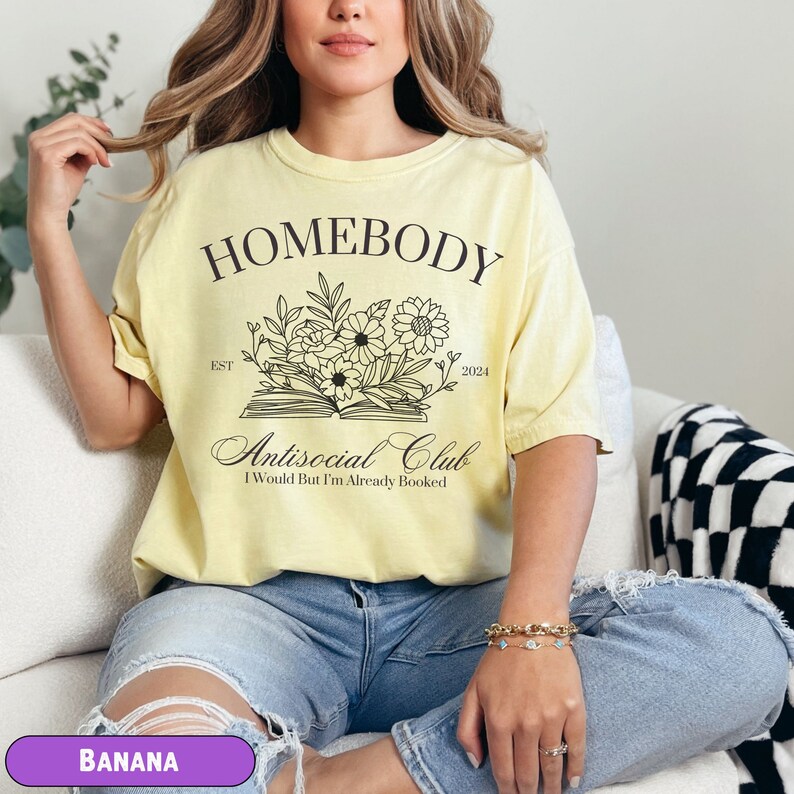 May include: A yellow t-shirt with the text "Homebody Est 2024 Antisocial Club I Would But I'm Already Booked" and a floral design.