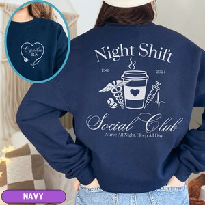 K&ouml;nnte beinhalten: Navyblauer Sweatshirt mit einer wei&szlig;en Grafik einer Kaffeetasse, einem Stethoskop, einer Pille und einer Spritze. Der Text "Night Shift Social Club" ist in einer kursiven Schrift geschrieben. Der Text "Nurse All Night, Sleep All Day" steht unter der Grafik. Der Text "EST 2024" steht &uuml;ber der Grafik.