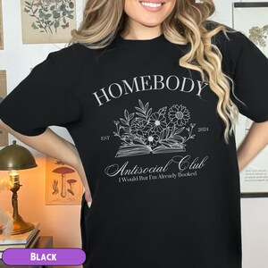 May include: Black t-shirt with white text that reads "Homebody Est. 2024 Antisocial Club I Would But I'm Already Booked". The text is surrounded by a floral design.