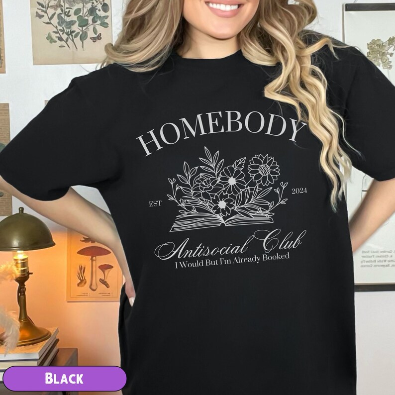 May include: Black t-shirt with white text that reads "Homebody Est. 2024 Antisocial Club I Would But I'm Already Booked". The text is surrounded by a floral design.