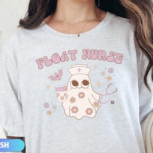 May include: Light gray t-shirt with the words "FLOAT NURSE" in pink bubble letters. The shirt features a cartoon ghost wearing a nurse's hat, holding a coffee cup, and a stethoscope. The ghost is decorated with flowers and a bat.