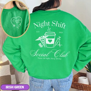 K&ouml;nnte beinhalten: Gr&uuml;ner Pullover mit einem wei&szlig;en Grafikdesign, auf dem "Night Shift Social Club" mit einer Kaffeetasse, einem Stethoskop, einer Spritze und einem Herzen steht. Der Text "EST" und "2024" befindet sich ebenfalls auf dem Pullover.