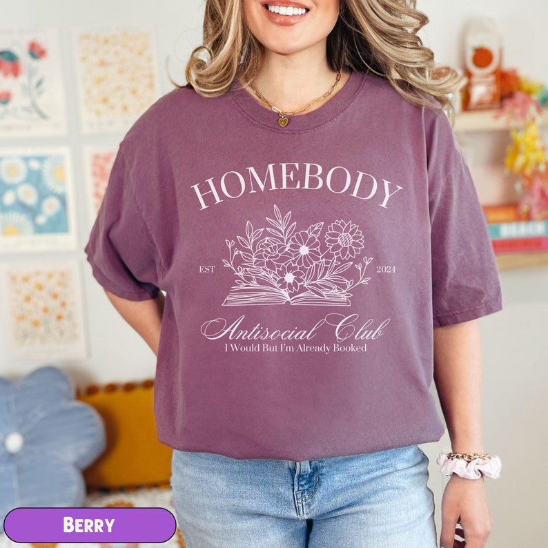 May include: A purple t-shirt with the text "Homebody Est. 2024" and "Antisocial Club I Would But I'm Already Booked" in white. The design features a book with flowers and leaves.