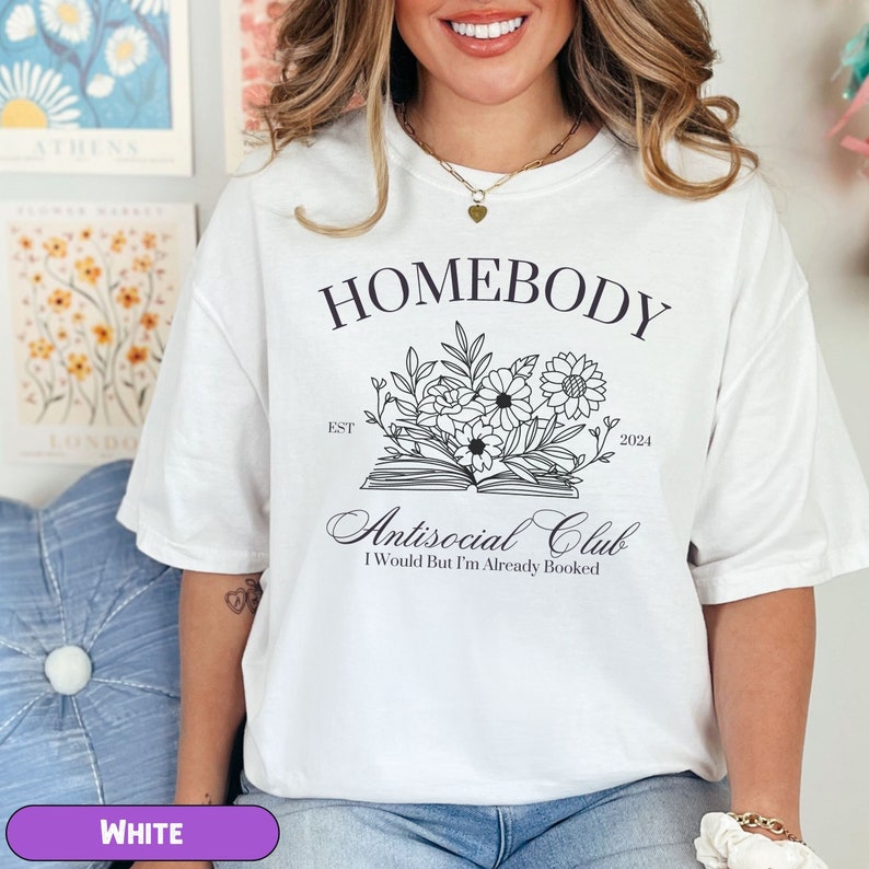 May include: White t-shirt with black text that reads "Homebody Est. 2024 Antisocial Club I Would But I'm Already Booked". The text is surrounded by a floral design with an open book.