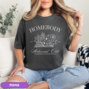 May include: A dark gray t-shirt with the text "Homebody Antisocial Club I Would But I'm Already Booked" and a floral design above the text. The text "Est 2024" is to the right of the floral design.