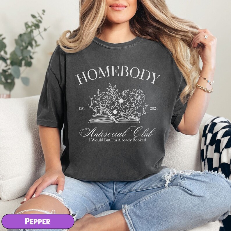 May include: A dark gray t-shirt with the text "Homebody Antisocial Club I Would But I'm Already Booked" and a floral design above the text. The text "Est 2024" is to the right of the floral design.