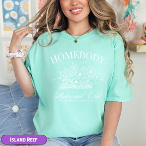 May include: A light green t-shirt with the text "Homebody Antisocial Club I Would But I'm Already Booked" in white. The shirt features a floral design and the text "Est. 2021".