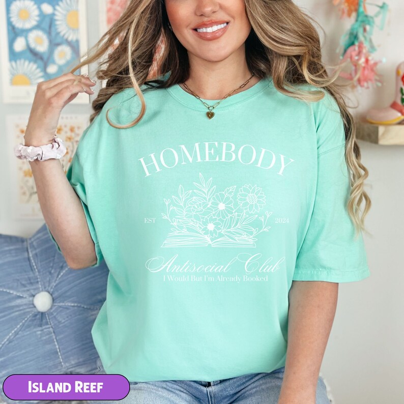 May include: A light green t-shirt with the text "Homebody Antisocial Club I Would But I'm Already Booked" in white. The shirt features a floral design and the text "Est. 2021".