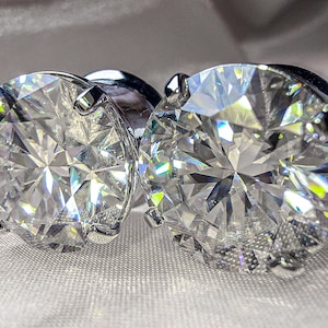 Könnte beinhalten: Zwei große, klare, runde, facettierte, diamantähnliche Steine, die in silberne Metallfassungen gefasst sind. Die Steine reflektieren das Licht und funkeln.