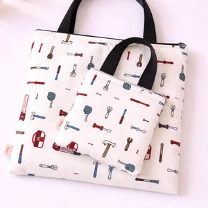 May include: Two white tote bags with black handles, one large and one small, both patterned with colorful tool illustrations. The tools include hammers, wrenches, and screwdrivers. The bags are made of fabric.