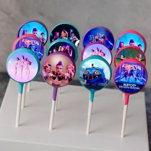 May include: A collection of round lollipops with colorful images and white sticks. The images feature animated characters and the text "KPOP DEMON HUNTERS". The lollipop sticks have teal, purple, and pink accents.