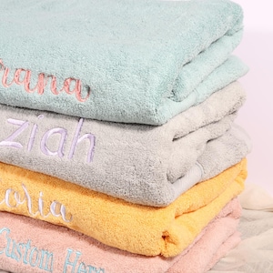 Op de afbeelding: Een stapel opgevouwen, gepersonaliseerde badhanddoeken in verschillende kleuren: mintgroen, grijs, geel en roze. Elke handdoek is geborduurd met een andere naam in een complementaire kleur. De handdoeken zijn zacht en pluizig.