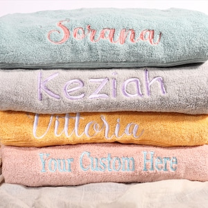 May include: Four pastel-coloured embroidered bath towels stacked on top of each other. The top towel is mint green with the name "Sorana" in pink. The next towel is grey with "Keziah" in purple. The third is yellow with "Vittoria" in white, and the bottom towel is pink with "Your Custom Here" in blue.