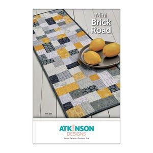 May include: A quilting pattern for a table runner called "Mini Brick Road" by Atkinson Designs. The pattern features a patchwork design with gray, black, and yellow squares. The pattern is shown on a table runner with lemons on a plate.